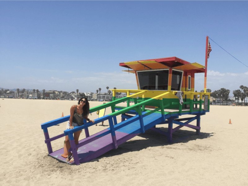 Abi standing at a multicoloured lifeguard hut on Venice Beach, LA.