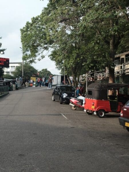 Street near a Sri Lankan train station with tuk-tuks, cars and people waiting by the road.