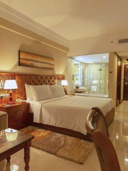 Large hotel room with king bed, lounge chair, desk and glass bathroom window.
